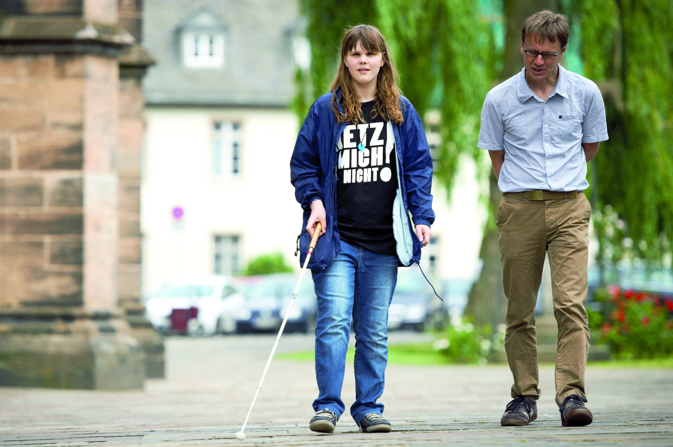 Szene aus dem Mobilitätsunterricht: Eine männliche Fachkraft für Blinden- und Sehbehindertenrehabilitation begleitet eine junge Frau mit Blindenlangstock. Sie trägt ein T-Shirt mit der Aufschrift "Hetz mich nicht!"